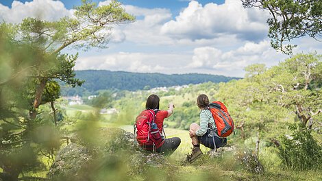 Zwei Wanderinnen mir Rucksäcken hocken auf Steinen und schauen in die Ferne.